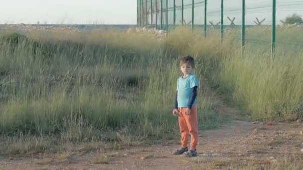 战争和武装冲突中的儿童 小男孩儿的破破烂烂的旧衣服站在边境的网状篱笆边 孤立无援儿童收容营移徙者 — 图库视频影像