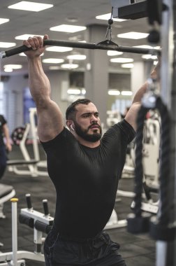 Spor salonunda ağırlık kaldırma antrenmanı. Siyah tişörtlü, ağırlık kaldıran genç sakallı bir adam.