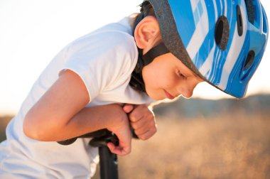 Yazın gün batımında açık hava oyunları sırasında scooterlı, mavi kasklı, şirin, mutlu beyaz çocuk.