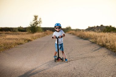 Güneş gözlüklü ve kasklı mutlu gülümseyen küçük çocuk Amerika Birleşik Devletleri 'ndeki boş çöl yolunda scooter standında duruyor.