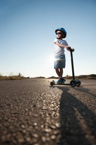 active sport little child in blue helmet and shorts wearing trendy sunglasses standing on asphalt road with mini scooter on hot sunset summer day