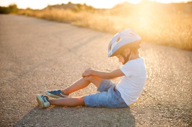 Spor sakatlığı konsepti. Kask takan küçük bir çocuk yaz günbatımında çölde asfalt yolda oturuyor.
