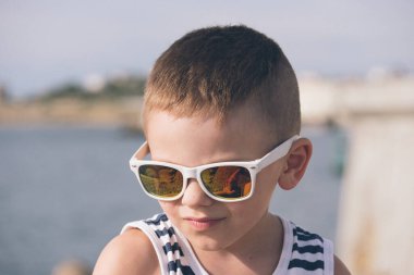 Gökyüzü ve denizin arka planında yelek ve güneş gözlüğü takmış küçük bir çocuğun portresi.