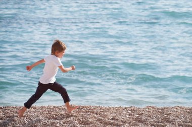 Deniz kıyısında sonbahar boyunca koşan beyaz gömlekli ve pantolonlu sağlıklı beyaz çocuk.
