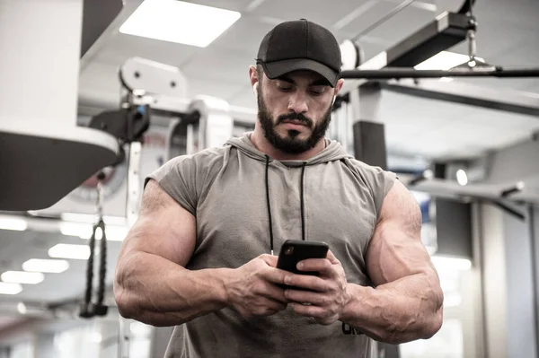 strong young bearded male with cap watch in cell phone smartphone during training pause
