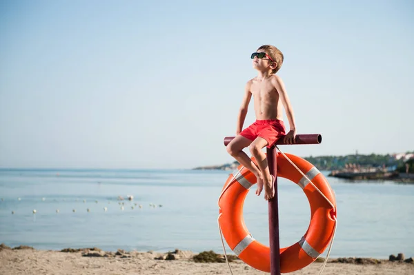 一个穿着红色短裤和太阳镜的小男孩 在晒黑的阳光下休闲活动时 坐在沙滩上 带着救生圈 — 图库照片