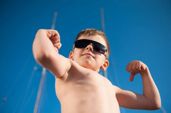 Copil puternic mic sport în ochelari de soare demonstrează mușchiul său mare în aer liber zi de vară — Fotografie, imagine de stoc