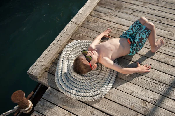 Sano bambino pigro in occhiali da sole e pantaloncini sdraiato sognando sul molo di legno con acqua di mare corda — Foto Stock