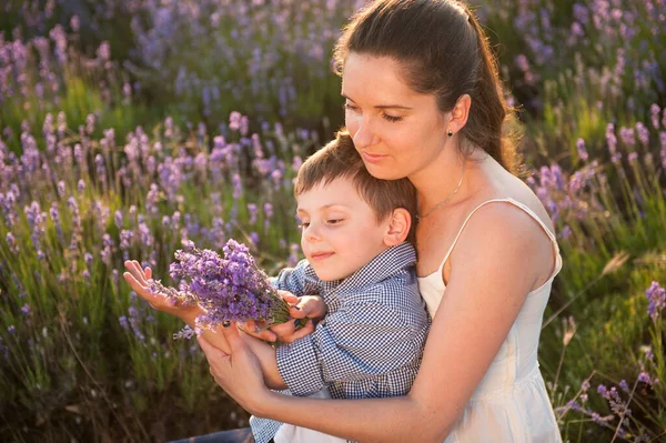 Adorable familia caucásica feliz joven madre con ramo de violeta y sonriente niño en la puesta del sol campo de flores de verano —  Fotos de Stock