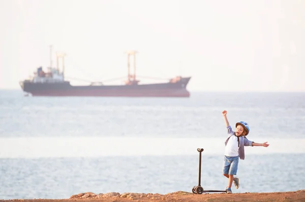 Χαρούμενο μικρό καυκάσιο παιδί σε κράνος με σκούτερ στη θάλασσα παραλία με φορτηγό πλοίο στον ορίζοντα — Φωτογραφία Αρχείου