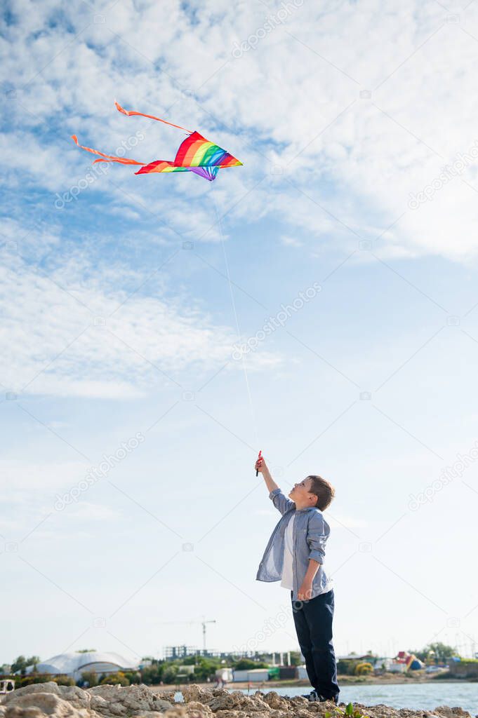 sport little caucasian boy with flying multicolored kite on blue sky background on summer day