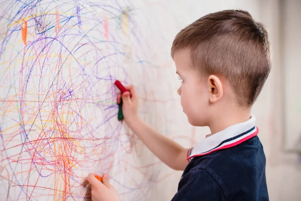 The child draws on the wall with a crayon. The boy is engaged in creativity at home