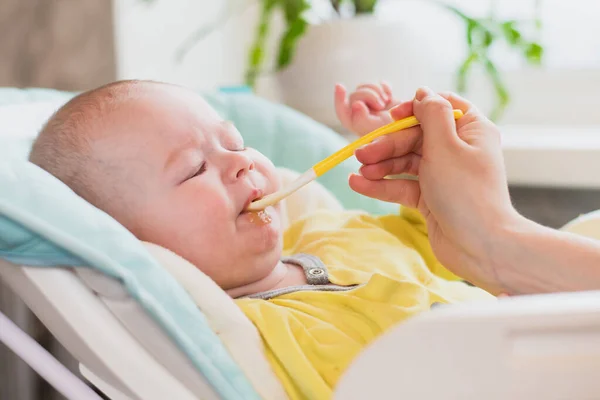 Mom feeds baby vegetable puree in a high chair. The mother puts a spoon of mashed in the child\'s mouth. The infant eats food.