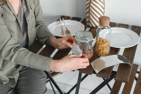 Una mujer pega una pegatina para la inscripción en un frasco de cereal. Organización del almacenamiento de cereales, pasta y otros productos en la cocina Fotos De Stock