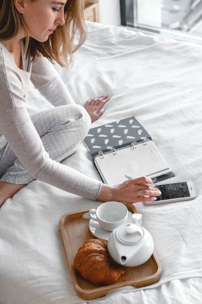 En kvinna planerar Affärer i en anteckningsbok och dricker kaffe, Frukost på sängen på morgonen. Kaffe och bakverk Stockbild