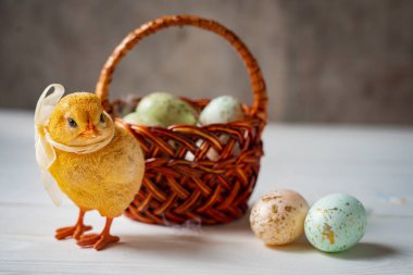 Paskalya dekoratif tavuğu beyaz ve gri ahşap arka planda bir sepet ve yumurtanın yanında duruyor..