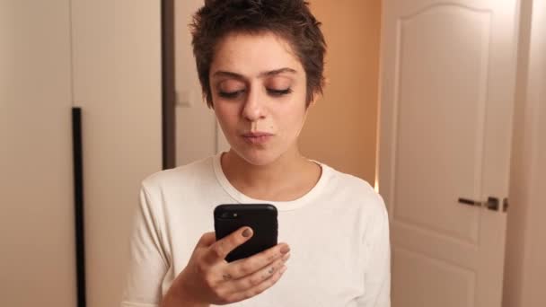Портрет коротковолосой девушки в белой футболке. Девушка нажимает на смартфон на фоне современного скандинавского стиля интерьера 4k — стоковое видео