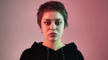 Kısa saçlı, neon ışınlı genç bir kızın portresi. Kız doğrudan kameraya bakıyor, saçına dokunuyor. 4k