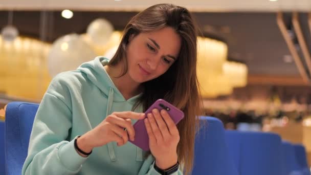 Cerca de la retrato de la mujer joven en una camisa de sudor turquesa se sienta en la cafetería y hace clic en un teléfono inteligente. Ella sonríe lindo 4k — Vídeos de Stock