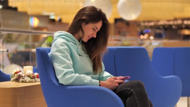 Portrait young woman in a turquoise sweatshirt. She sits in a duty free and clicks a smartphone. She expects her flight — Stock Video