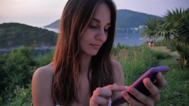 Ritratto di ragazza sullo sfondo di montagne, oceano, Paesaggio esotico. Lei fa clic sui pulsanti del suo smartphone. Concetto di vacanza 4k — Video Stock