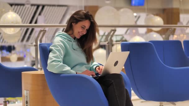 Mujer joven se sienta en el vestíbulo y mira a la pantalla del ordenador portátil. Ella sonríe y se alegra 4k — Vídeos de Stock