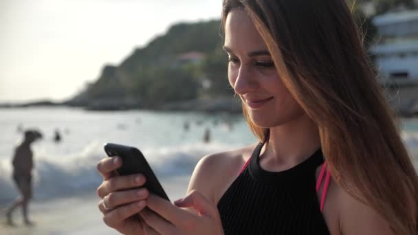 Портрет молодой девушки. Она стоит на берегу океана, улыбается и печатает что-то по телефону. 4k — стоковое видео