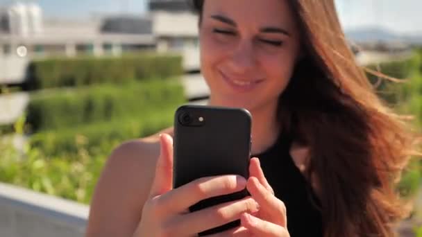 Portrait en gros plan de souriante jeune femme élégante bissneswoman lavec les yeux marron profond en robe noire essayant d'acheter quelque chose d'agréable avec son smartphone fond urbain de la ville. Mouvement lent 4k — Video