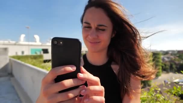 Primo piano ritratto di giovane bissneswoman elegante sorridente lwith occhi marroni profondi in abito nero cercando di acquistare qualcosa di piacevole con il suo smartphone sfondo urbano della città. Rallentatore 4k — Video Stock