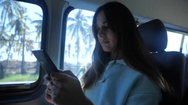 Een jonge vrouw leest een elektronisch boek in een bus. In het raam een zonnige dag, palmbomen op de achtergrond 4k — Stockvideo