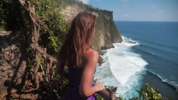 Retrato cerrado de una joven sonriente con pelo marrón, el viento sopla, mirando al mar. Chica en una camisa de sudor turquesa caminando sobre las rocas. Movimiento lento 4k — Vídeos de Stock