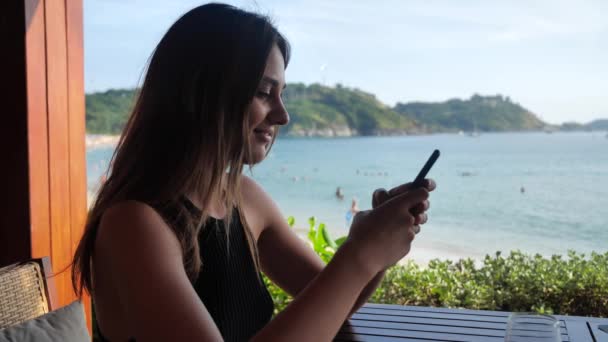 Une jeune fille s'assoit dans un kiosque et clique au téléphone. Elle sourit doucement, discute avec quelqu'un sur le fond de la plage 4k — Video