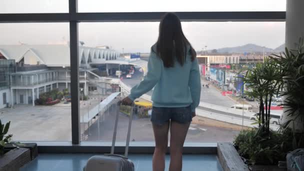 Giovane ragazza sta aspettando il suo volo vicino alla finestra in aeroporto con bagaglio 4k — Video Stock