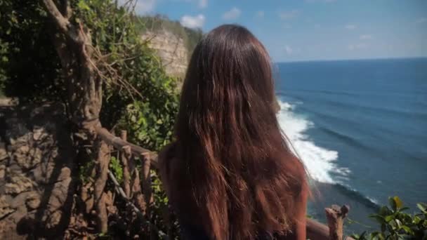Retrato de close-up de uma jovem mulher sorrindo com cabelos castanhos, o vento sopra, olhando para o oceano. Menina em uma camisola turquesa viajando sobre as rochas. Movimento lento 4k — Vídeo de Stock