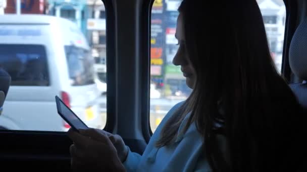 Una joven está leyendo un libro electrónico en un autobús. En la ventana un día soleado, palmeras en el fondo 4k. — Vídeo de stock