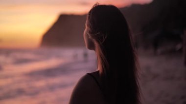 Sunset above ocean. Young woman standing on a beach her wind blows on her hair 4k