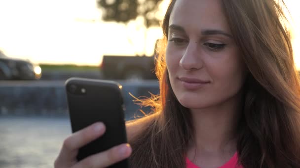 Gros plan de sourire jeune fille élégante et attrayante avec les yeux marron profond en robe noire en essayant d'acheter quelque chose d'agréable avec son smartphone fond urbain de la ville.Mouvement lent 4k — Video