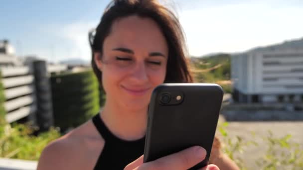 Γκρο πλαν πορτρέτο της χαμογελαστή νεαρή κομψή bissneswoman lμε βαθιά καστανά μάτια σε μαύρο φόρεμα προσπαθεί να αγοράσει κάτι ευχάριστο με το smartphone Urban φόντο της πόλης. Αργή κίνηση 4k — Αρχείο Βίντεο