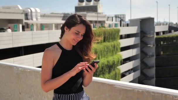 Closeup του χαμογελαστού νεαρό κομψό ελκυστικό κορίτσι με βαθιά καστανά μάτια σε μαύρο φόρεμα προσπαθεί να αγοράσει κάτι ευχάριστο με το smartphone Αστικό φόντο της πόλης.Αργή κίνηση 4k — Αρχείο Βίντεο