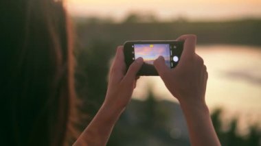 Güzel deniz günbatımının fotoğraflarını ve videolarını çeken genç bir kadının dikiz görüntüsü. Çekici kadın gün batımında deniz kıyısında fotoğraf çekiyor. Gün batımında sahilde fotoğraf çeken bir kadın. 4K