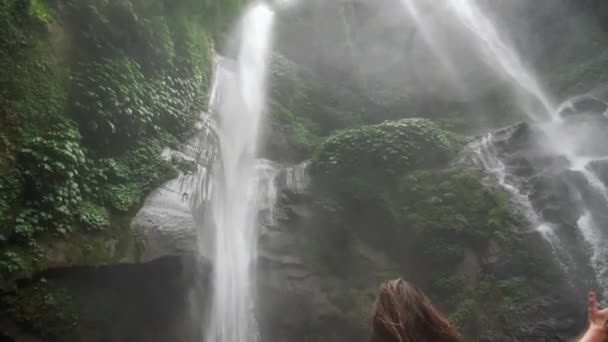 Aantrekkelijke vrouw open armen spectaculaire waterval in Indonesië. Schot van een jonge vrouw die haar armen uitstrekt bij een majestueuze waterval. Succes- en prestatieconcept 4k — Stockvideo