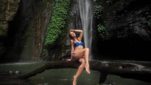 Belle femme assise sur une bûche au bord du lac dans une forêt tropicale sur un fond de cascade sous la pluie. Unité avec la nature 4k — Video