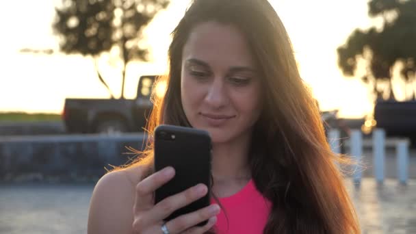 Közelkép portré gyönyörű lány mosolyog, és kattintson az okostelefon. Város háttér, naplemente 4k — Stock videók