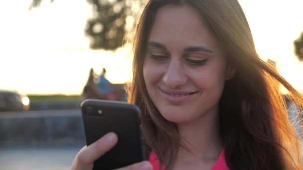 Menina bonita sorri e clica no smartphone. Fundo da cidade, por do sol 4k — Vídeo de Stock