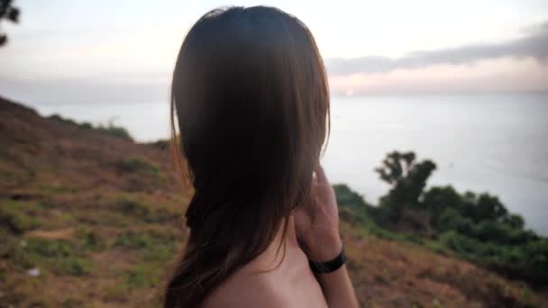 Κοντινό πορτραίτο ενός κοριτσιού που στέκεται πάνω από ένα γκρεμό και κοιτάζει τον ωκεανό. Τα μαλλιά της είναι άνεμος. φόντο ηλιοβασιλέματος 4k — Αρχείο Βίντεο