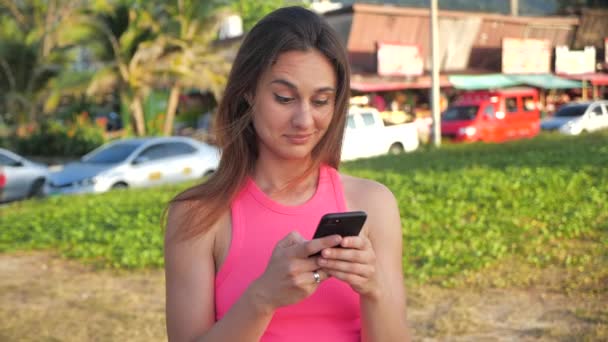 Retrato de una chica atractiva con una camisa corta rosa. Ella está de pie en las palmas del sitio en el fondo. Cute es condenado, sosteniendo un teléfono inteligente en sus manos. 4k — Vídeos de Stock