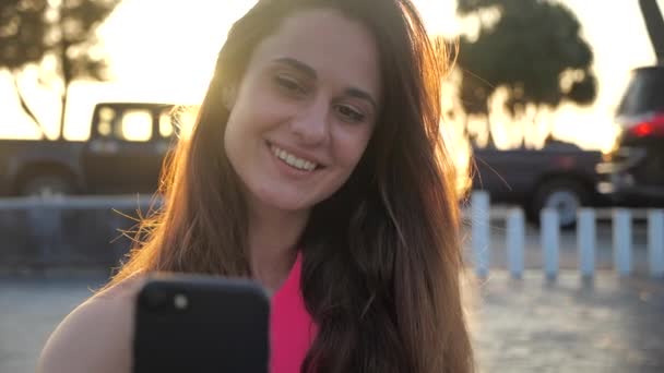 Nahaufnahme Porträt der schönen Mädchen lächelt und klickt auf das Smartphone. Weibliche Blicke in die Kamera. Hintergrund der Stadt, Sonnenuntergang 4k — Stockvideo