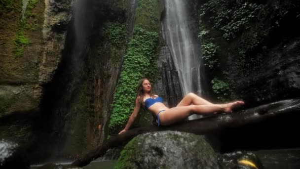 Krásná žena sedí v plavkách u jezera v deštném pralese na pozadí vodopádu v dešti. Jednota s přírodou 4k — Stock video