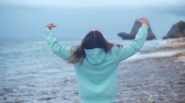 Güzel genç bir kadının portresi yaklaşır rüzgar gibi kollarını kaldırır, saçlarını savurur ve günbatımında huzurlu bir sahil kenarında eğlenir.