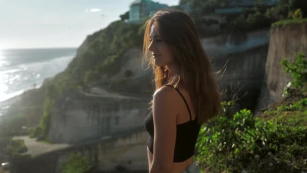 Portrét dívky, která stojí nad útesem a dívá se na oceán. Vlasy jí foukají větrem. Pozadí západu slunce 4k — Stock video